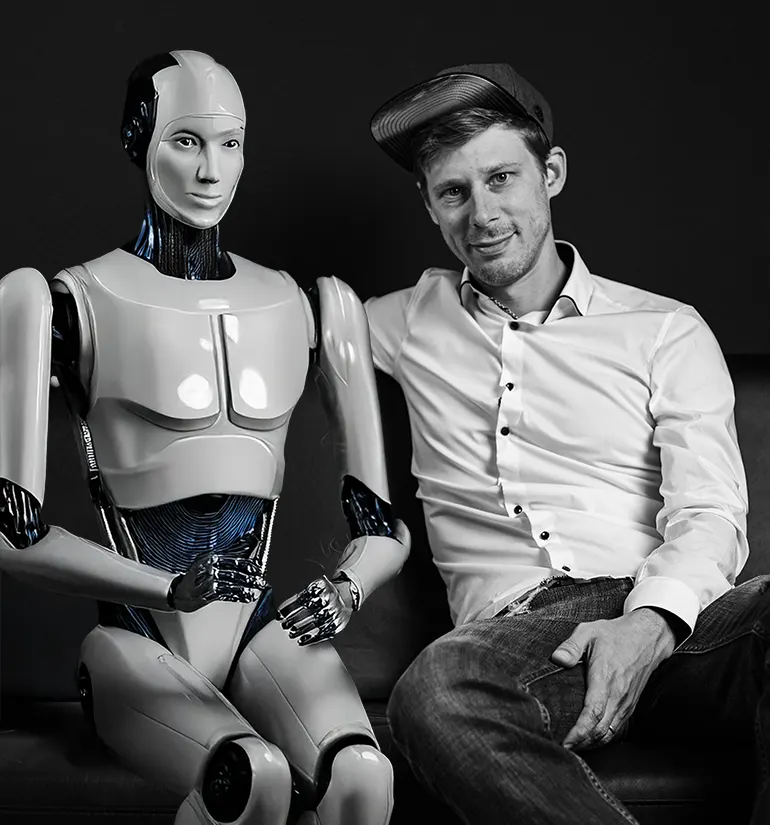 KI Webdesigner aus Freiburg sitzt mit humanoidem Roboter auf dem Sofa